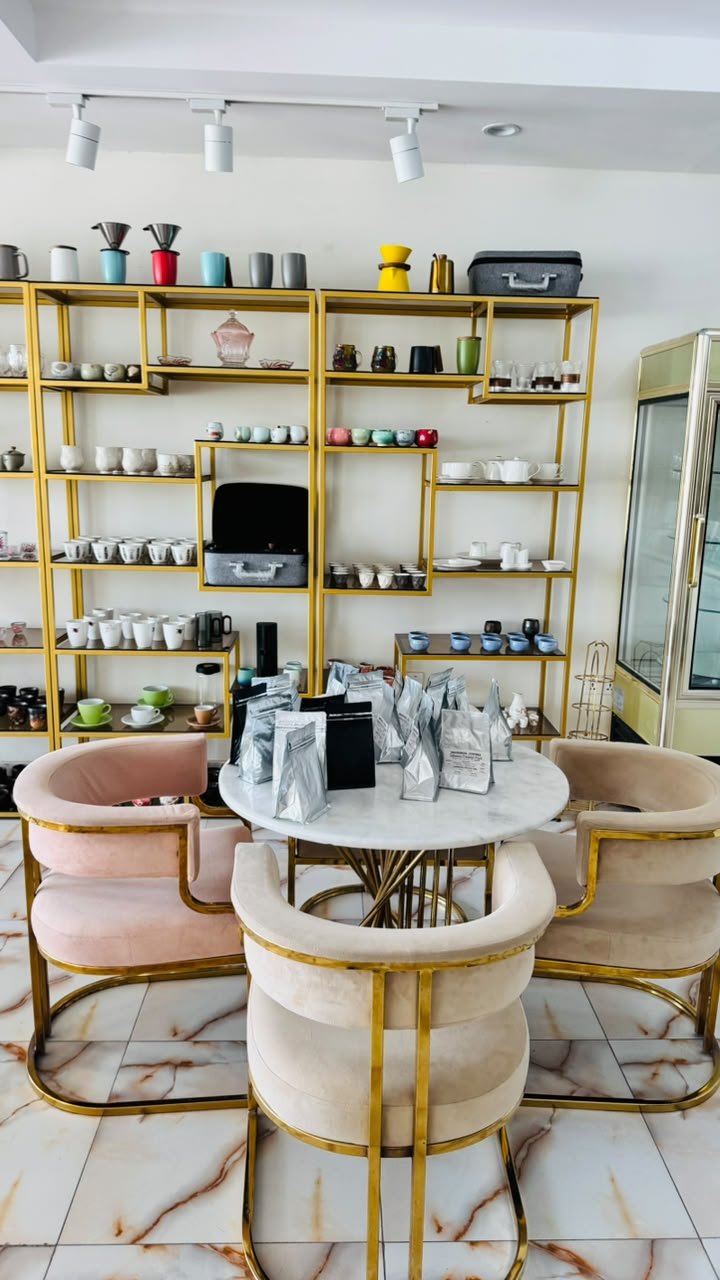 Café seating area with a round marble table, blush and cream velvet chairs with gold frames, and gold-framed shelving displaying cups and coffee bags