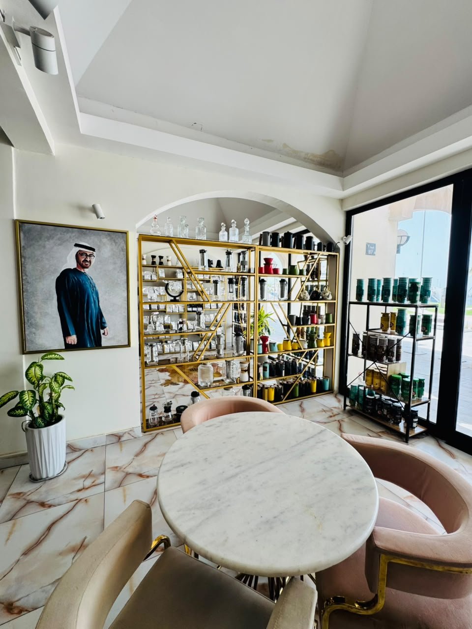 Dining corner with a round marble table and pink velvet chairs in front of an arched alcove with gold shelving, beside a large window